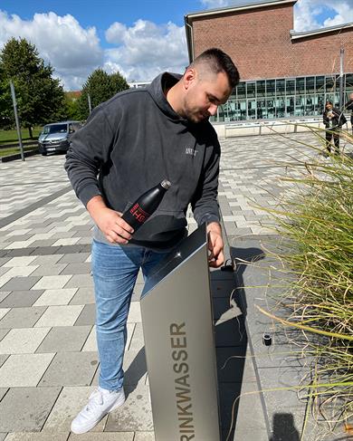 Ein junger Mann steht vor einem Trinkbrunnen und drückt den Aktivierungsknopf auf der Rückseite des Geräts, um den Wasserfluss zu starten.